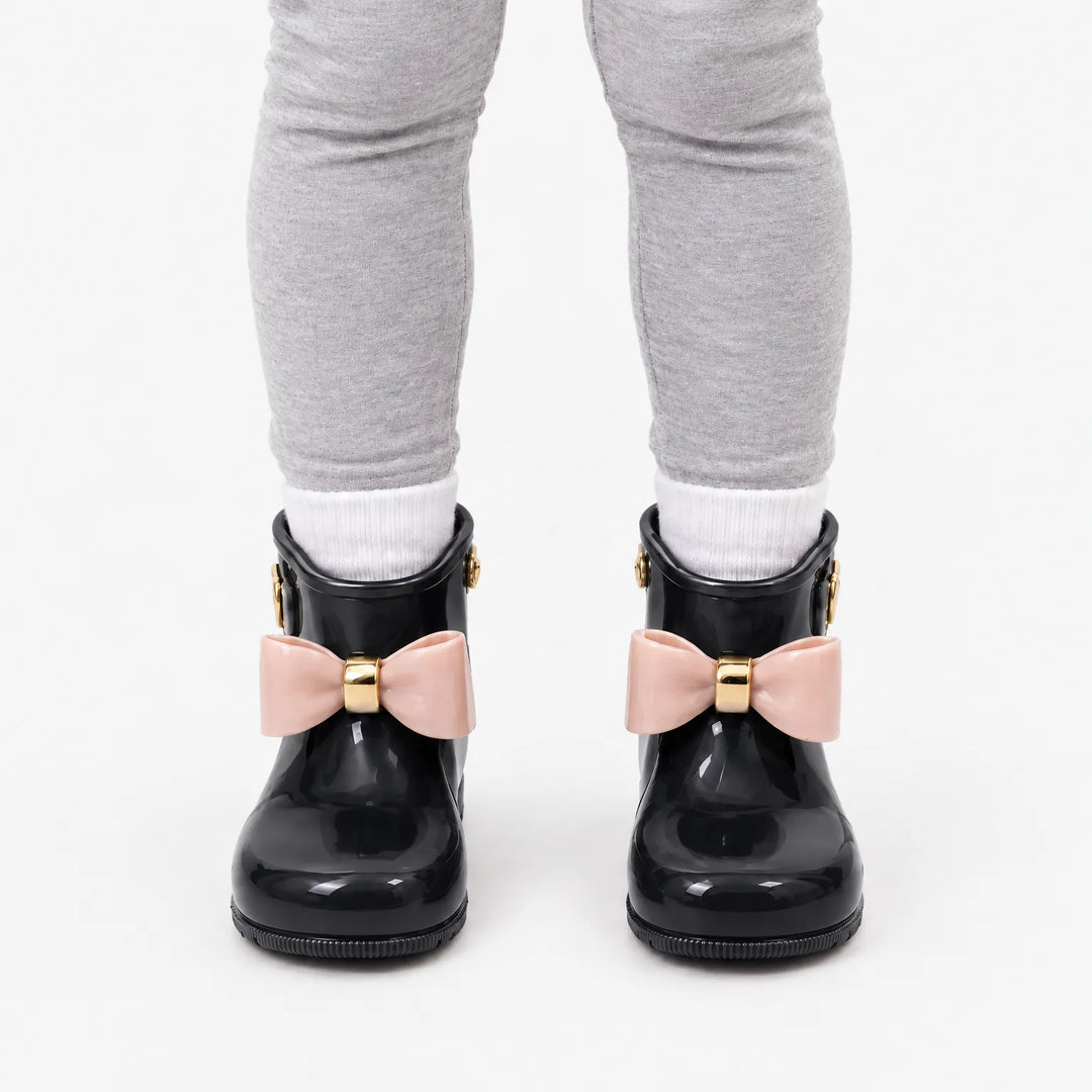 Front view of a child in grey leggings and white socks wearing black wellies with contrasting pink bows and gold bands. 9