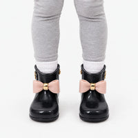 Front view of a child in grey leggings and white socks wearing black wellies with contrasting pink bows and gold bands. 9