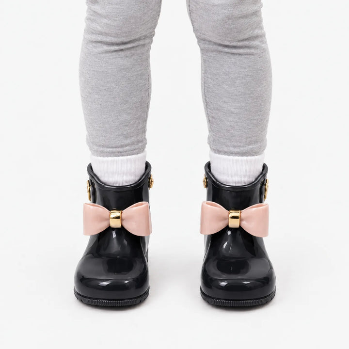 Front view of a child in grey leggings and white socks wearing black wellies with contrasting pink bows and gold bands. 9