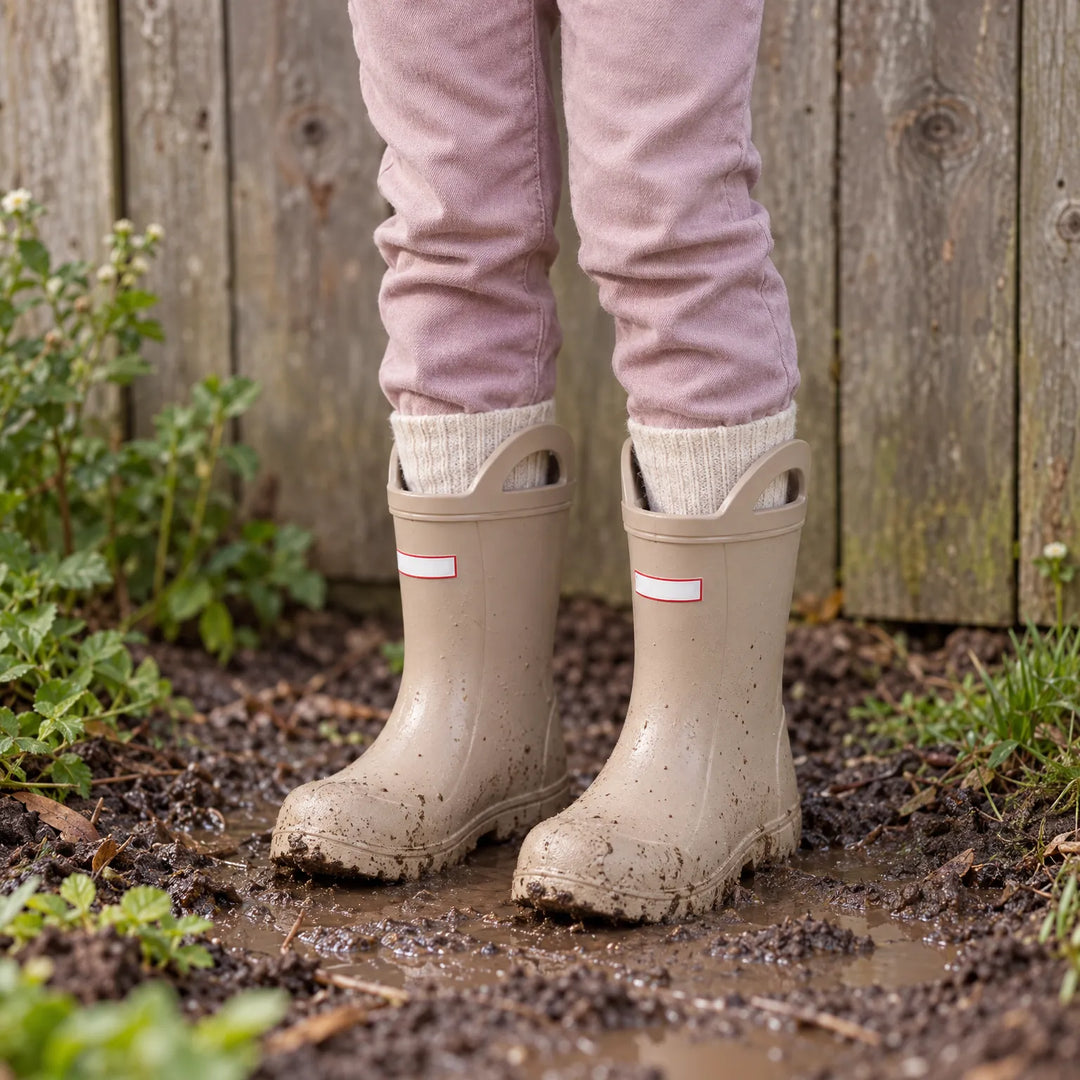 Child wearing muddy beige wellies with handles over cream socks, standing in a muddy garden patch by a wooden fence. 3