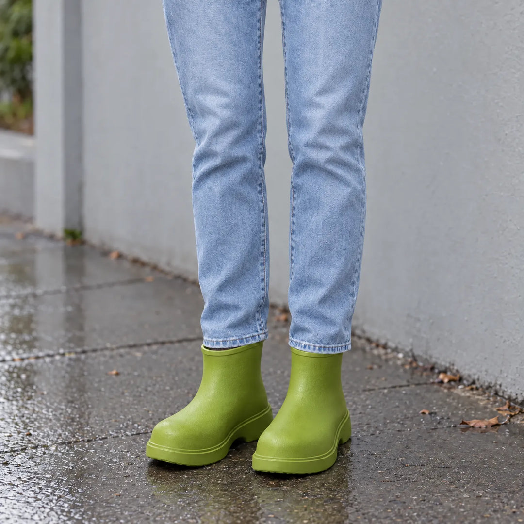 Green ankle boots worn with light-wash jeans on a wet pavement beside a grey wall, showing the rainy-day look. 1