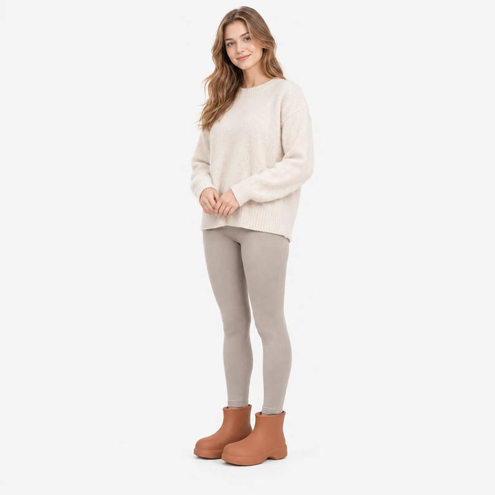 Full-length view of a smiling model in a cream knit sweater and grey leggings wearing rust-brown ankle boots. 4