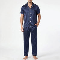Front view of a model in navy satin pyjama set with white piping, one hand in trouser pocket, on plain background. 2
