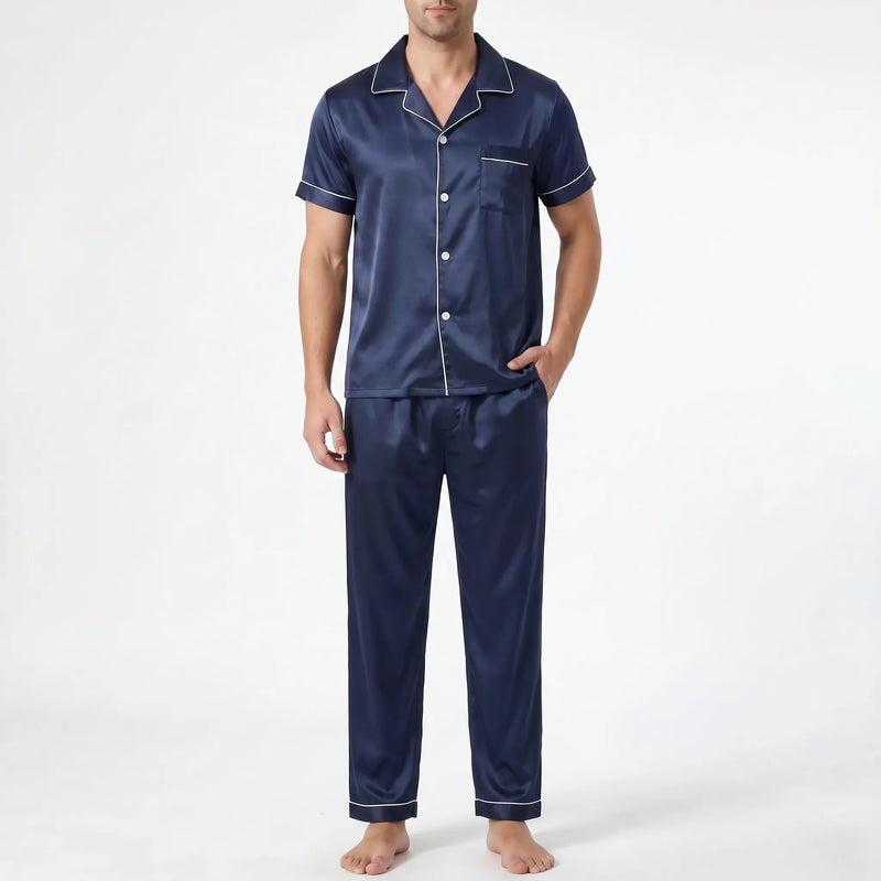 Front view of a model in navy satin pyjama set with white piping, one hand in trouser pocket, on plain background. 2