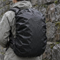Hiker in a grey rain jacket seen from behind, wearing a black rucksack cover beaded with raindrops against a wet stone wall. 1