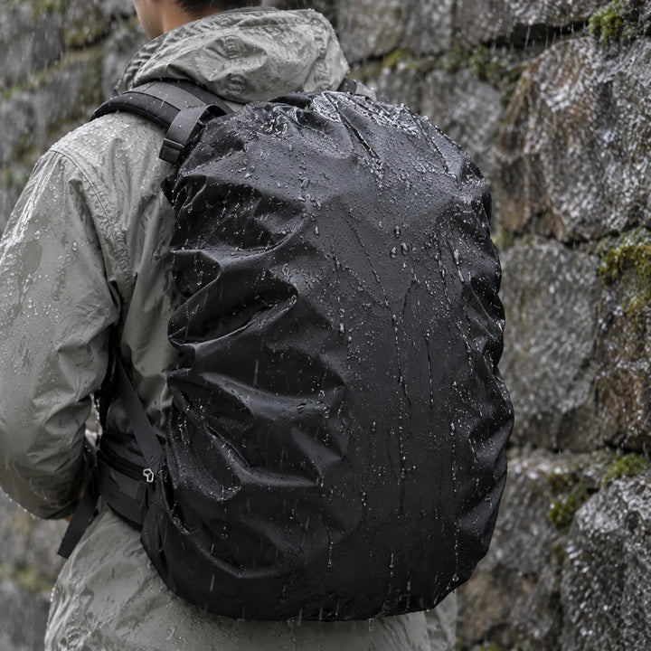 Hiker in a grey rain jacket seen from behind, wearing a black rucksack cover beaded with raindrops against a wet stone wall. 1