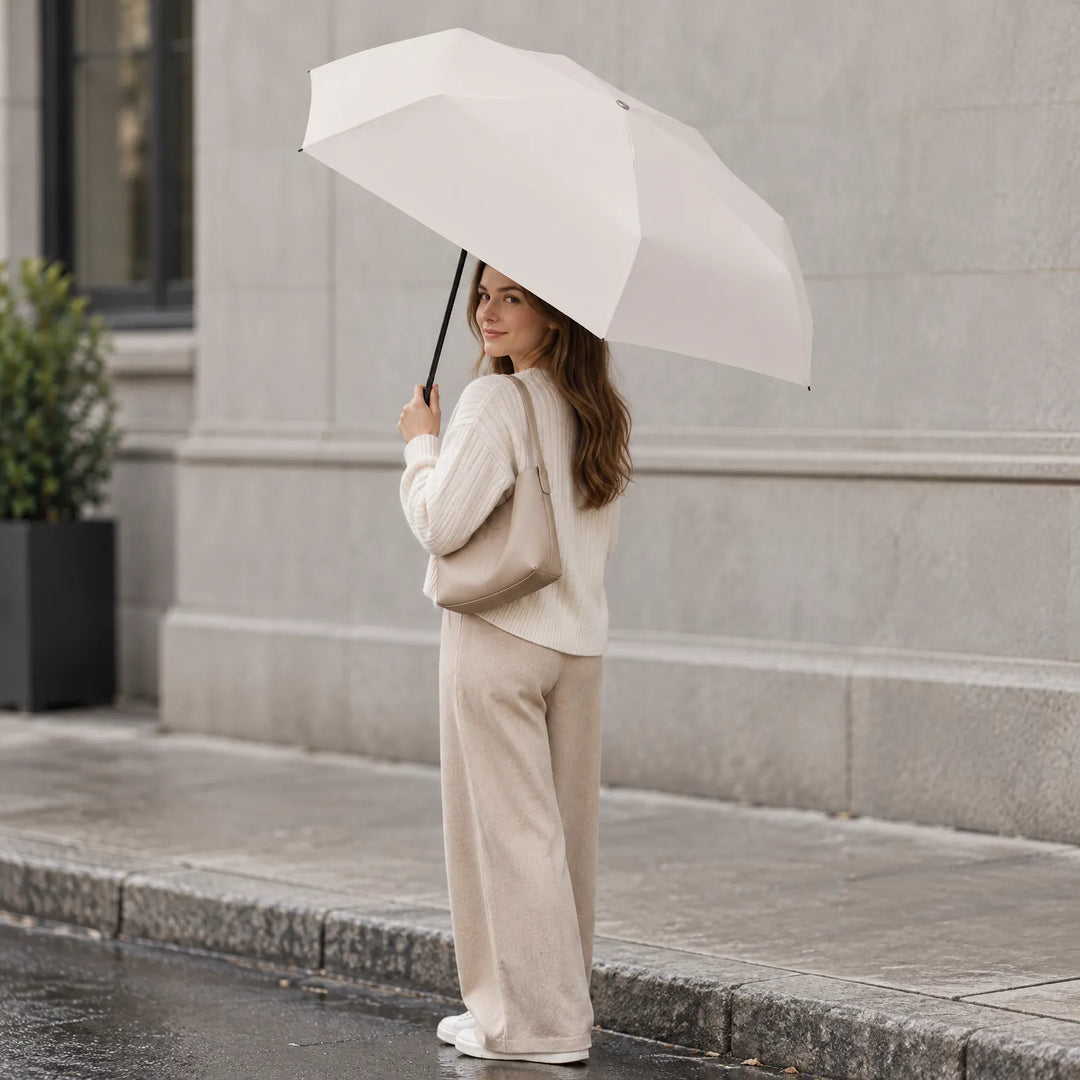 Woman in a cream knit sweater and beige trousers holds an open cream umbrella on a wet city pavement, smiling over her shoulder. 1