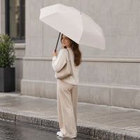Woman in a cream knit sweater and beige trousers holds an open cream umbrella on a wet city pavement, smiling over her shoulder. 1