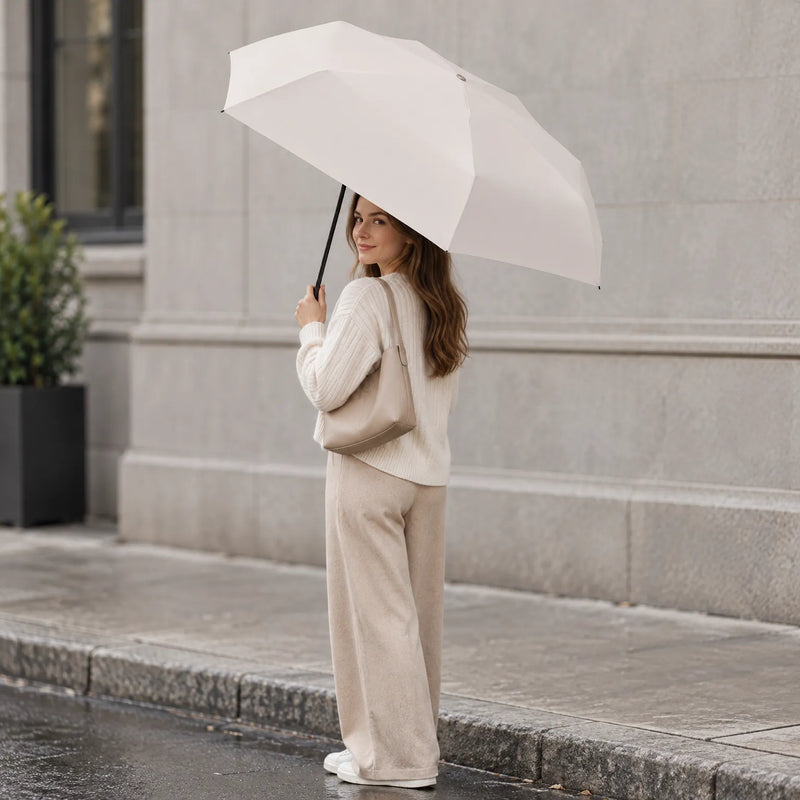 Woman in a cream knit sweater and beige trousers holds an open cream umbrella on a wet city pavement, smiling over her shoulder. 1