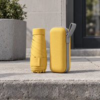 Mustard yellow folded umbrella standing next to its matching hard zip case with grey loop, set on a concrete step outdoors. 3