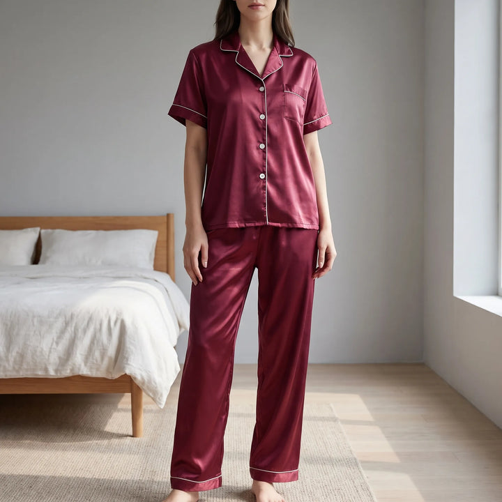 Model wearing the burgundy satin short-sleeve pyjama set with white piping, standing in a bright bedroom beside a wooden bed. 1