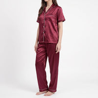 Three-quarter side view of a model in the burgundy satin pyjamas, showing the notched collar, pocket and straight-leg trousers. 4