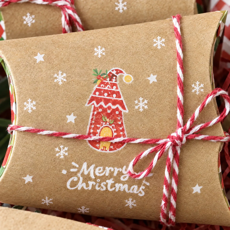 Close-up of a kraft pillow box with a red gingerbread-style house design and snowflakes, wrapped in red and white twine tied in a bow. 3