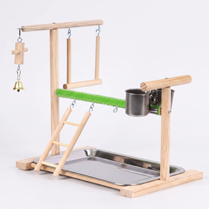 Wooden bird playstand on a plain background, seen from the front left, with ladder, swing, bell, green perch, steel cups and tray. 1