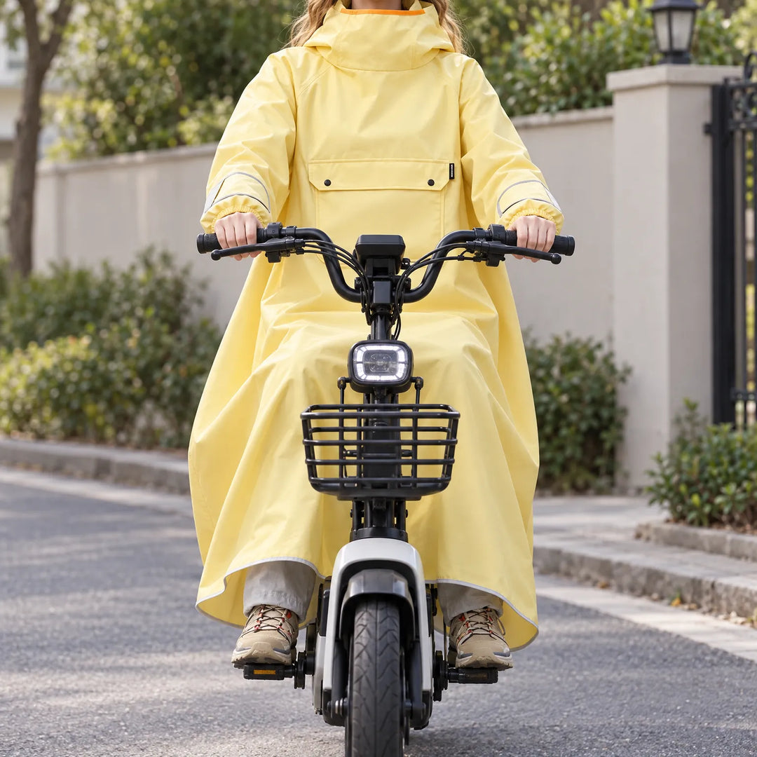 Rider in a pale yellow hooded poncho sits on a white e-scooter with a front basket, the cape draping over the legs. 1