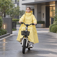 Smiling woman riding a white electric bike with a basket through a modern courtyard, wearing the yellow poncho with the hood up. 3