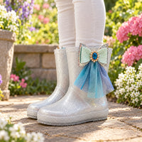 Child wearing silver glitter rain boots with a jewelled mint bow and blue tulle streamers, standing on a garden path among flowers. 1