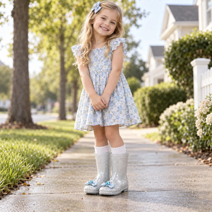 Smiling girl in a blue floral dress and white socks wearing silver glitter boots with small blue crown-and-bow charms on a sidewalk. 3
