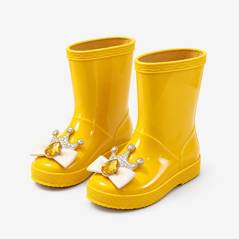 Pair of glossy yellow rain boots at an angle, each with a gold crown, cream bow and amber teardrop gem on the toe. 5