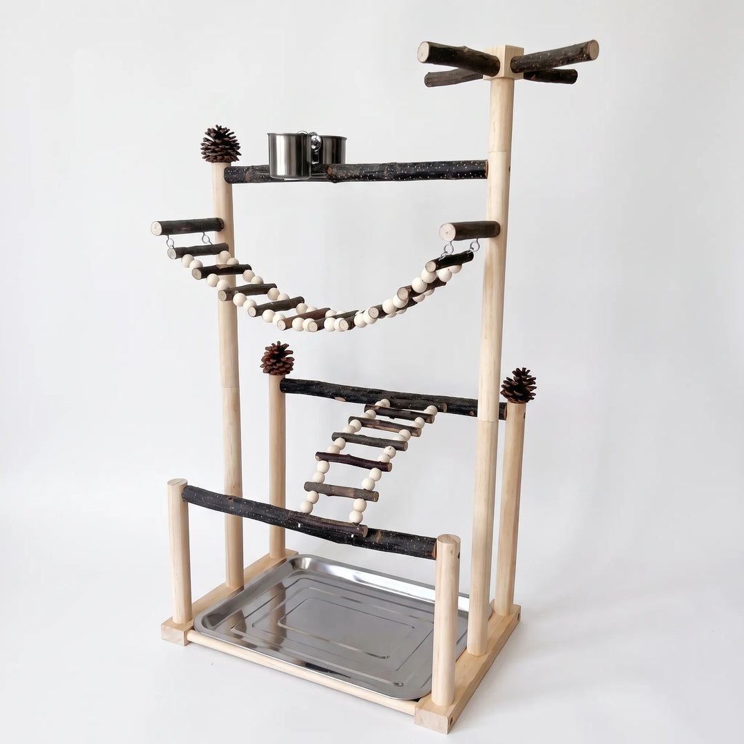 Wooden bird playstand on a plain background, with natural branch perches, a bead rope bridge, a ladder, pine cones, a steel food cup and a tray. 1