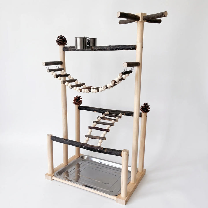 Wooden bird playstand on a plain background, with natural branch perches, a bead rope bridge, a ladder, pine cones, a steel food cup and a tray. 1