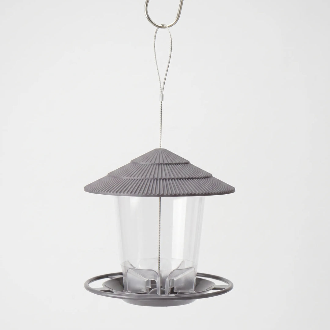 Grey feeder with ribbed tiered roof and clear seed tube, hanging from a steel wire loop and hook against a plain backdrop. 1
