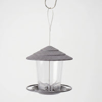 Grey feeder with ribbed tiered roof and clear seed tube, hanging from a steel wire loop and hook against a plain backdrop. 1