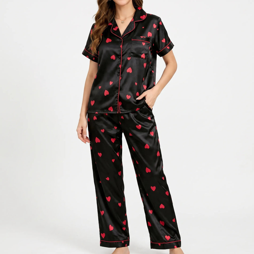 Front view of a model standing against a plain white background in the black satin pyjama set, one hand in a trouser pocket. 2