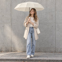 Smiling woman in a beige coat and jeans holds the open cream umbrella overhead while standing by a concrete wall. 1