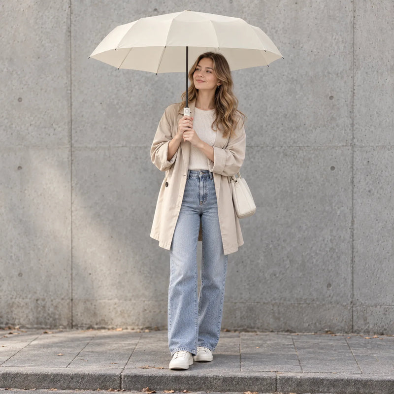 Smiling woman in a beige coat and jeans holds the open cream umbrella overhead while standing by a concrete wall. 1