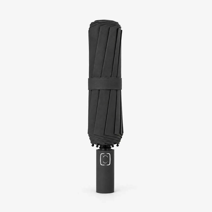 Folded black umbrella standing upright on a white background, wrapped with a fabric strap and showing its square handle emblem. 2