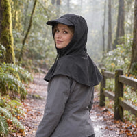 Woman in a black hooded cape with beaded raindrops, worn over a grey jacket on a damp forest trail. 3