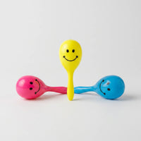 Three glossy plastic maraca rattles with smiley faces in yellow, pink and blue, one standing upright between two lying down, on a plain background. 1