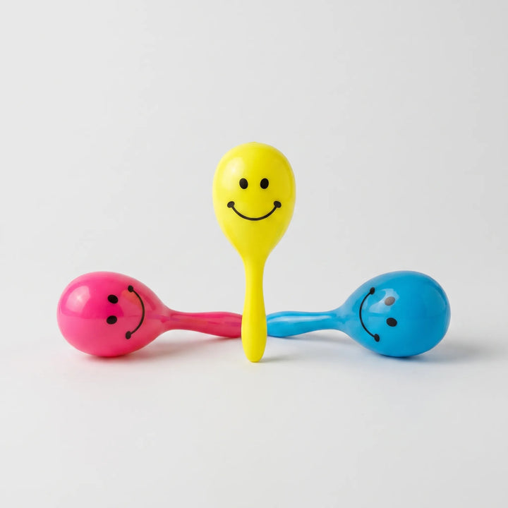 Three glossy plastic maraca rattles with smiley faces in yellow, pink and blue, one standing upright between two lying down, on a plain background. 1