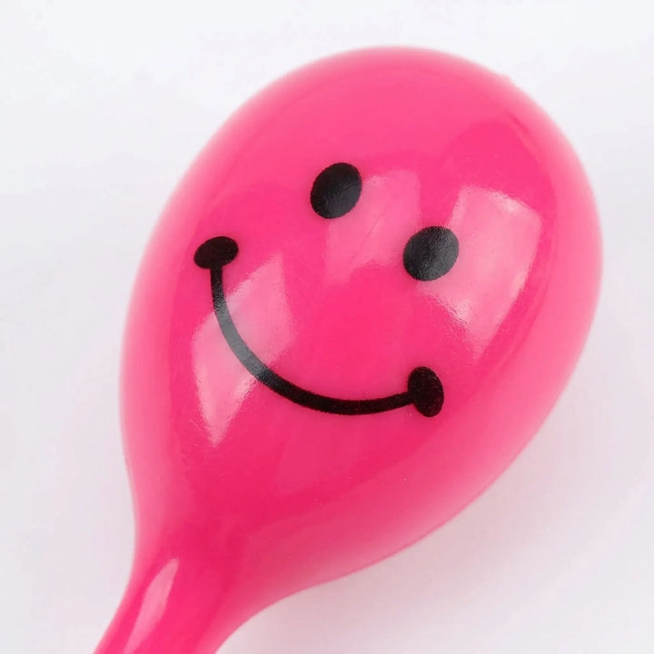 Close-up of the head of a glossy pink maraca rattle, showing its printed black smiley face and smooth plastic finish. 3