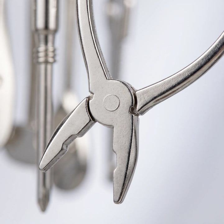 Close-up of the miniature silver pliers charm, showing the pivot and jaws, with other tool charms blurred behind. 3