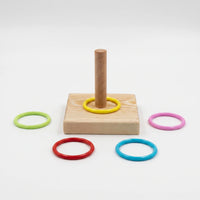 Wooden square base with an upright peg, one yellow ring on it and green, pink, red and blue rings laid around on white. 1
