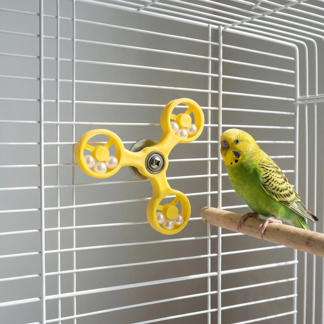 Yellow spinner toy attached to a white cage, with a green budgerigar perched on a wooden perch next to it. 3