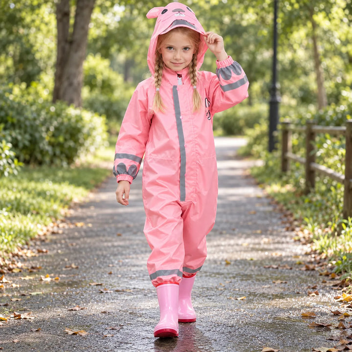 Smiling girl with braids walking along a park path in the pink rain jumpsuit with bunny-ear hood, lifting the hood brim. 3