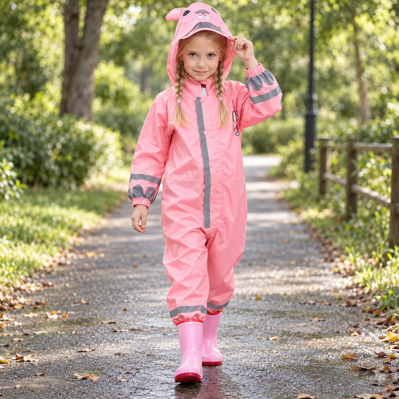 Smiling girl with braids walking along a park path in the pink rain jumpsuit with bunny-ear hood, lifting the hood brim. 3