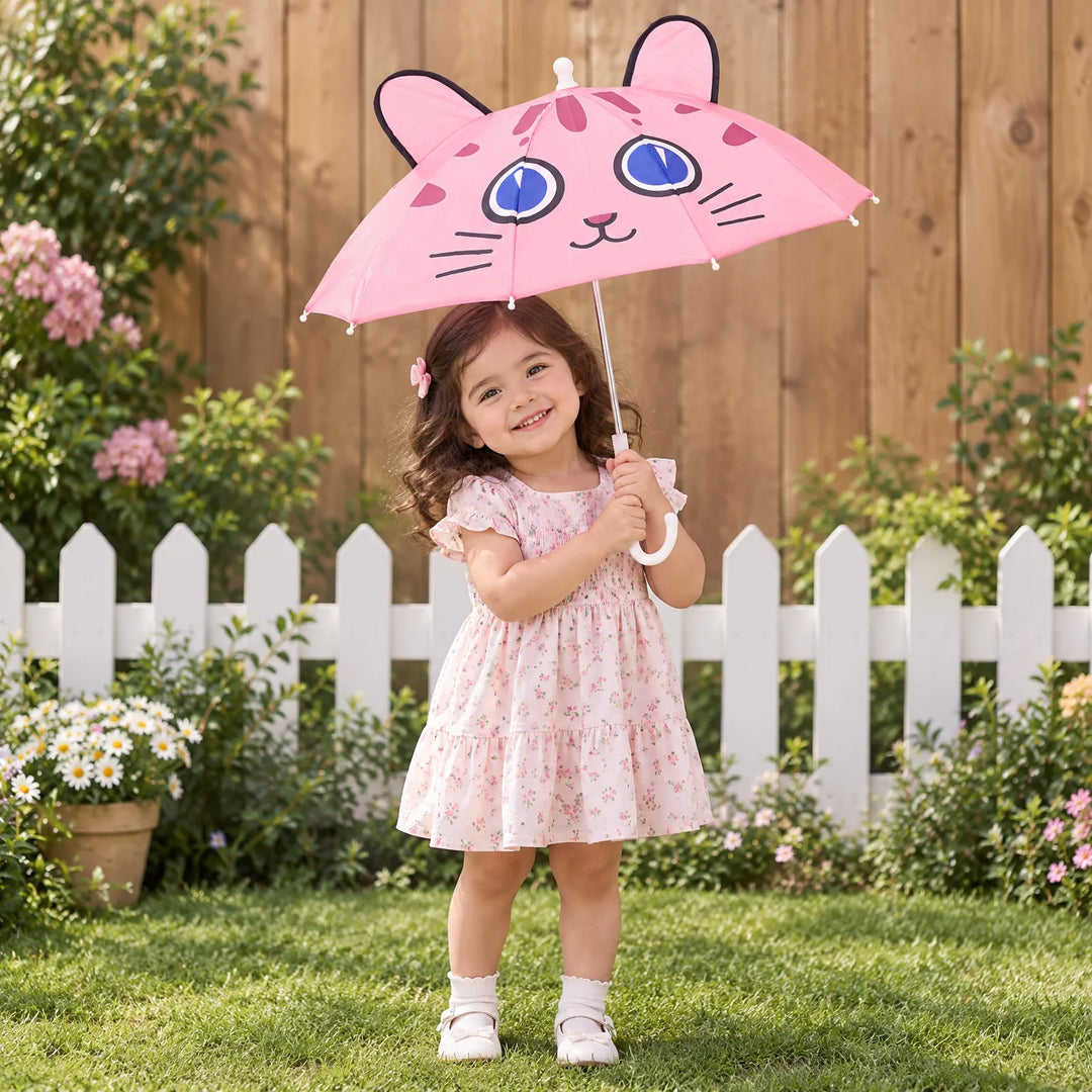 Smiling toddler in a floral dress holds the pink cat-face umbrella with ears in a sunny garden by a white fence. 1