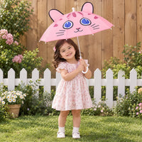 Smiling toddler in a floral dress holds the pink cat-face umbrella with ears in a sunny garden by a white fence. 1