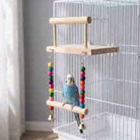 Blue and white budgie perched on the swing beneath the wooden platform, hung inside a white cage with colourful beads and bells. 2