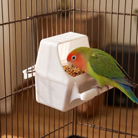 Green lovebird with a red face perched on the feeder fixed to the cage bars, eating seed from the tray. 2