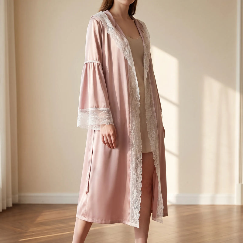 Model in a sunlit room wears the open pink satin robe with white lace edging and flared lace-cuffed sleeves. 1