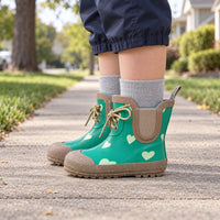 Child standing on a pavement in green heart-print rubber boots with tan toe caps, laces, elastic side panels, grey socks and navy trousers. 1