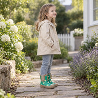 Smiling girl in a cream hooded jacket and jeans stands on a stone garden path wearing the green heart-print boots. 3