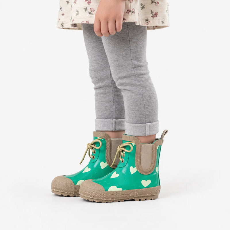 Side view of a child in grey leggings and a floral dress wearing the green heart boots against a plain white background. 4