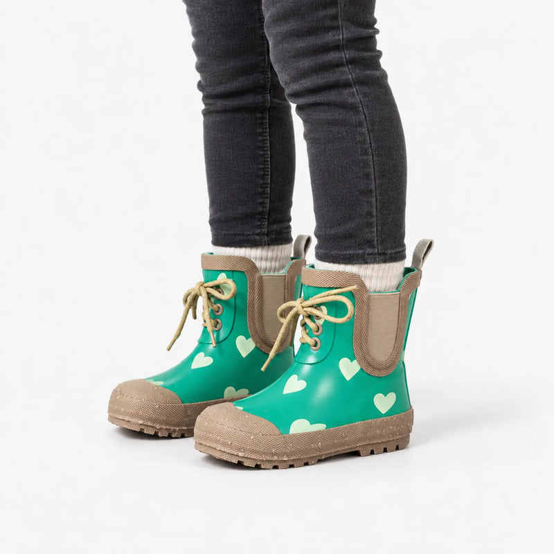 Child in dark grey jeans and cream socks wearing the green heart-print lace-up boots, seen from the side on white. 5