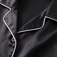 Close-up of the notched collar of the black satin pyjama shirt, with white piping, stitching and a grey button. 5